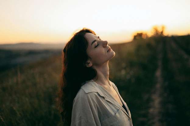 A woman with closed eyes stands in a field at sunset, appearing serene and contemplative.
