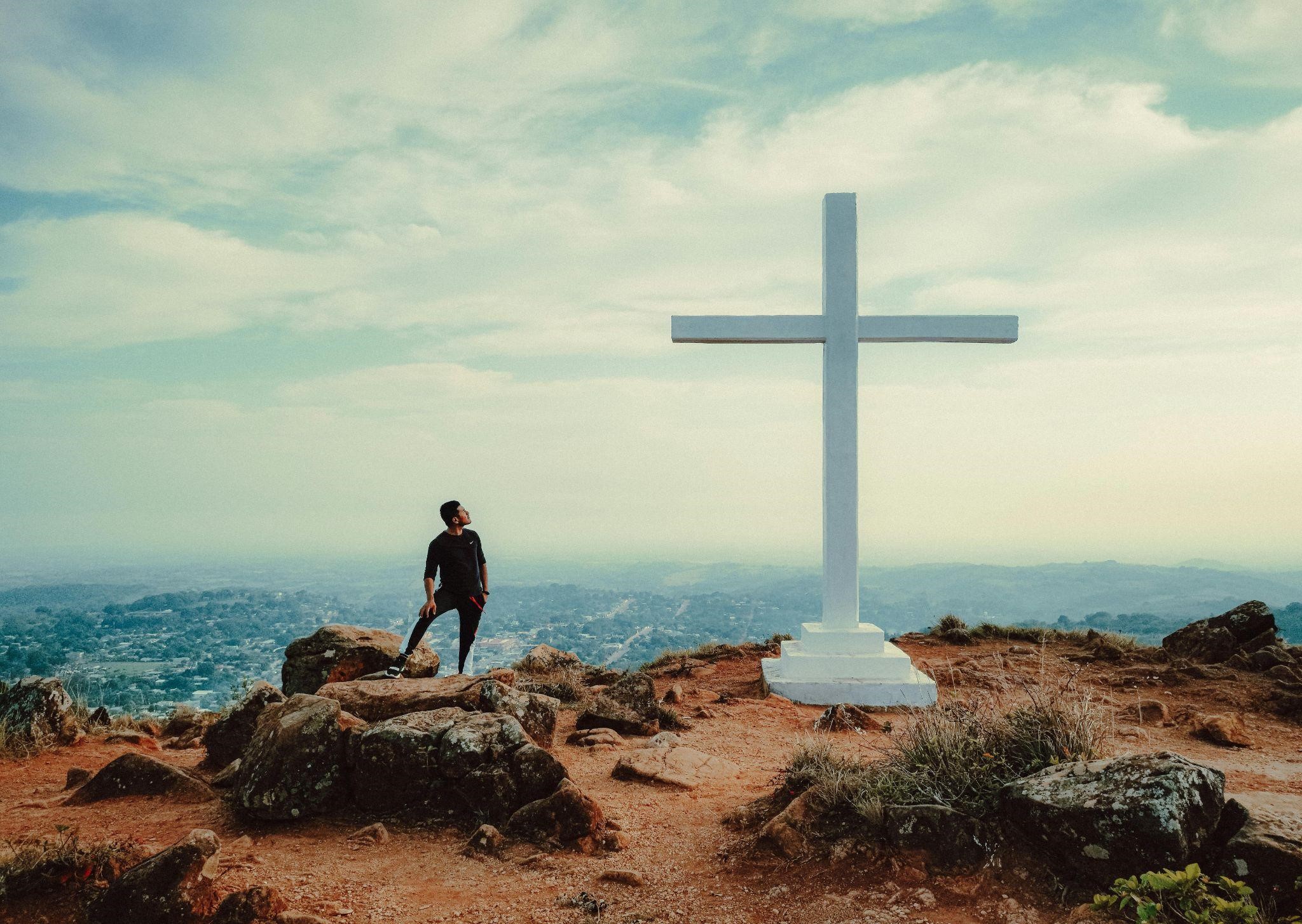 A person stands on a rocky hilltop beside a large cross, conveying the ways to stand firm in your faith.